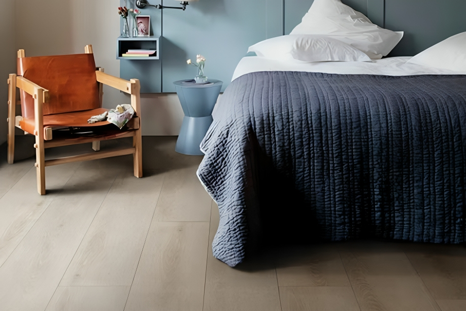 En harmonisk soveromsinnredning som reflekterer nåværende gulvtrender med et elegant lyst treparkettgulv, kombinert med en dypblå sengesprei og en cognacfarget lenestol som tilfører en varm og sofistikert atmosfære.